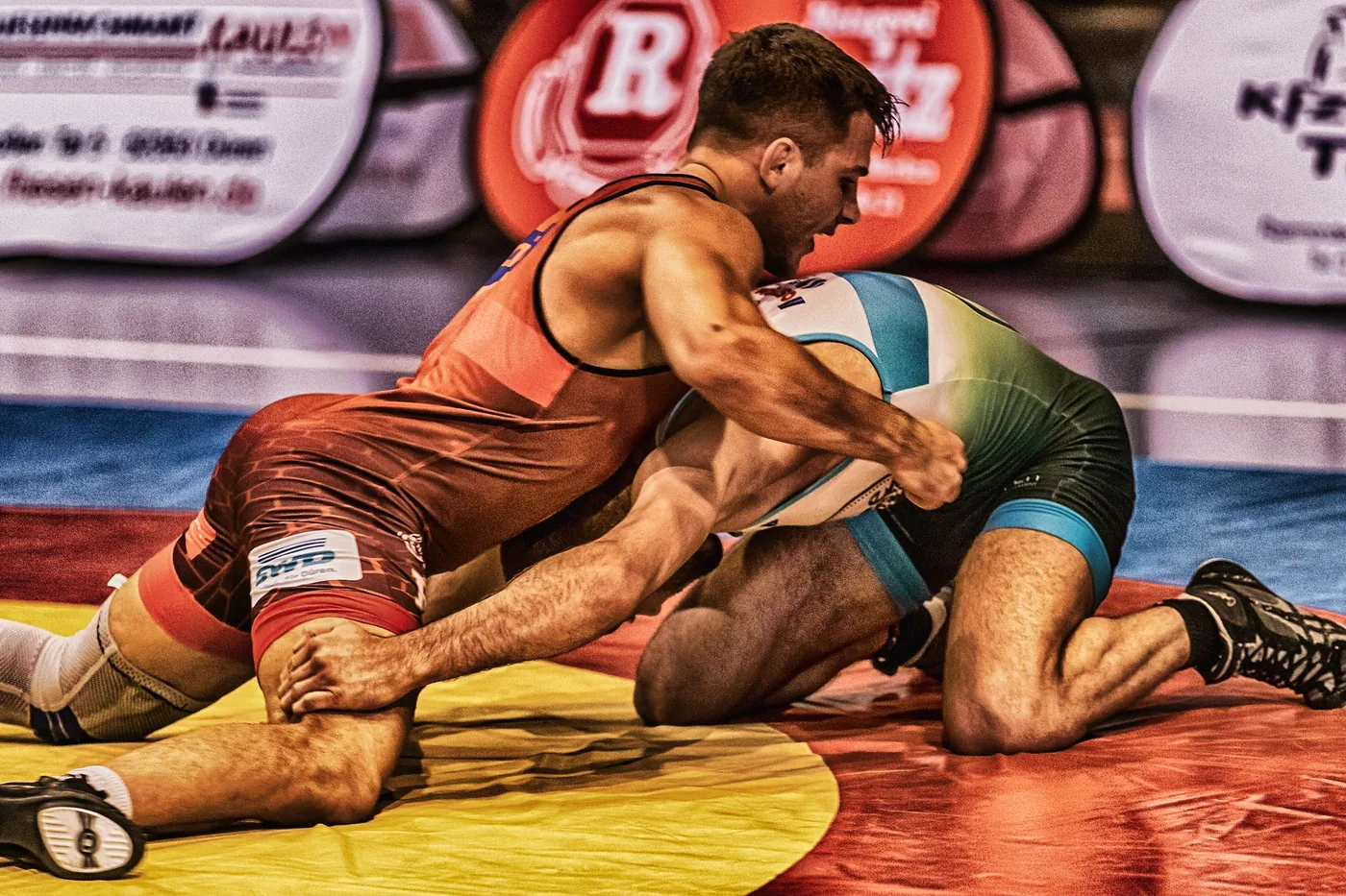 Two wrestlers grapple on a red-and-yellow mat in a gym, one in maroon pinning a teal-and-green opponent, with blurred signs behind.