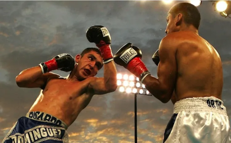 Two boxers trade punches in a ring at dusk under bright lights, one in blue trunks labeled DOMINGUEZ.