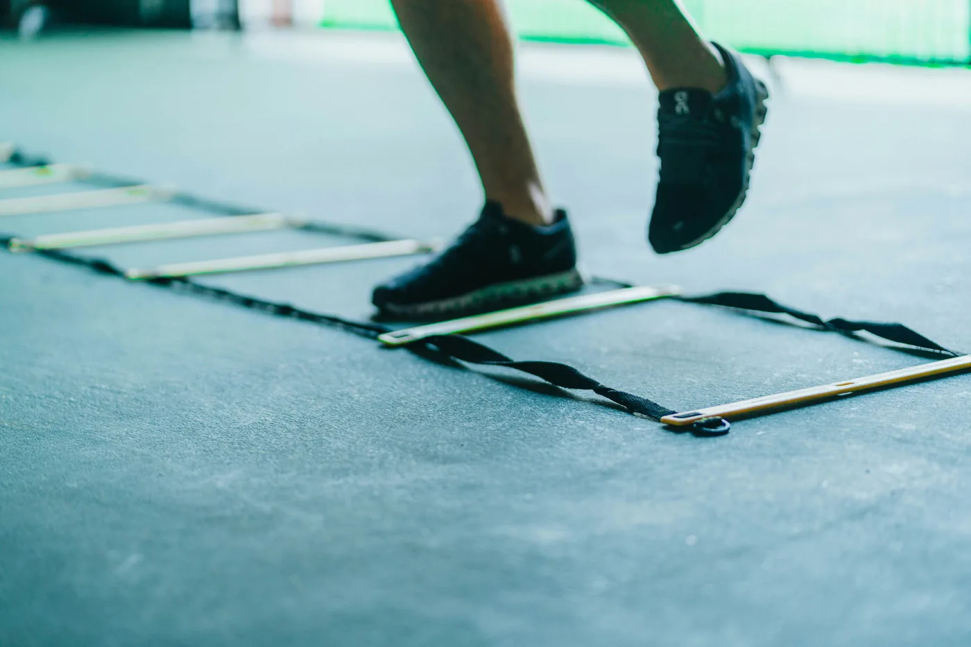 Close-up of a person’s legs and black sneakers stepping through an agility ladder on a gym floor during training