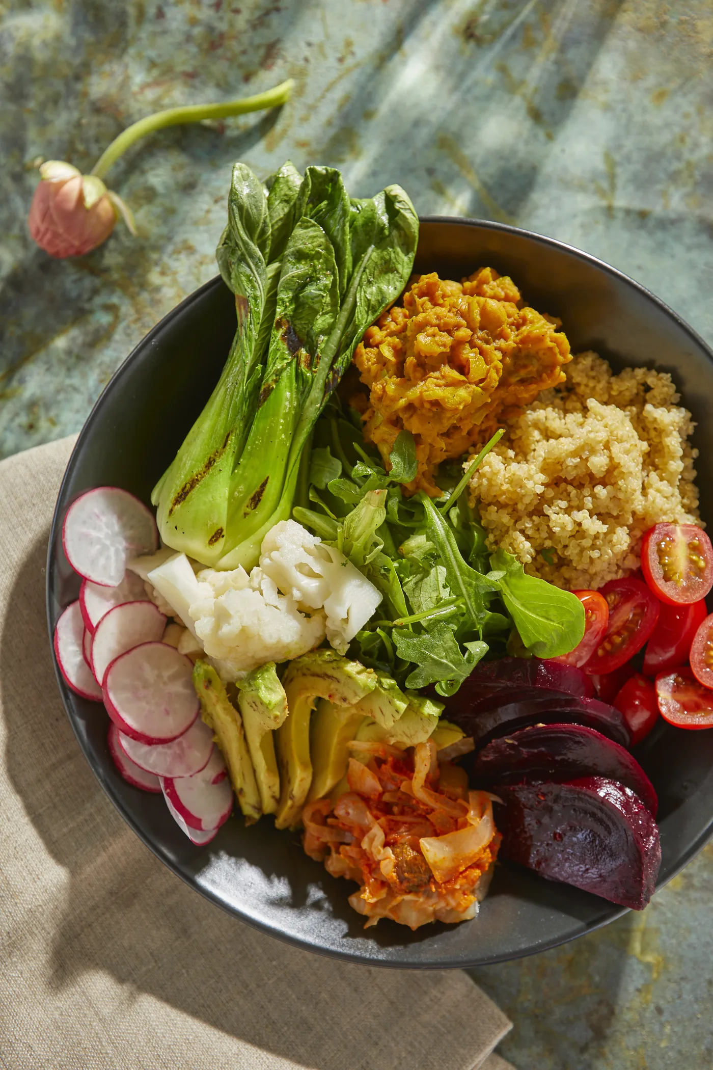 Colorful veggie bowl with bok choy, quinoa, radishes, beets, tomatoes, avocado, cauliflower and kimchi on a sunlit table