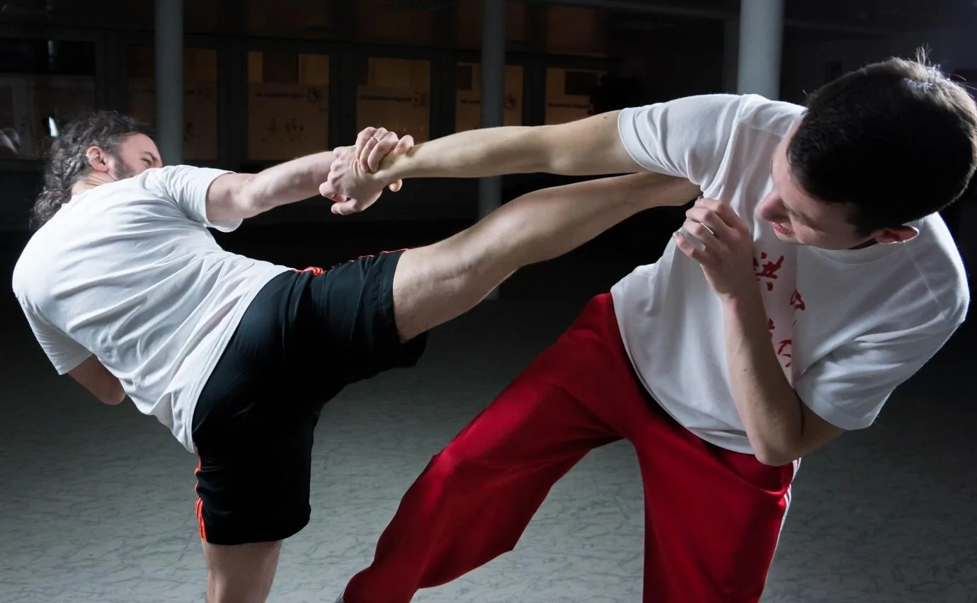 Two men spar in martial arts practice, one delivering a high kick while the other blocks in a dim indoor studio.