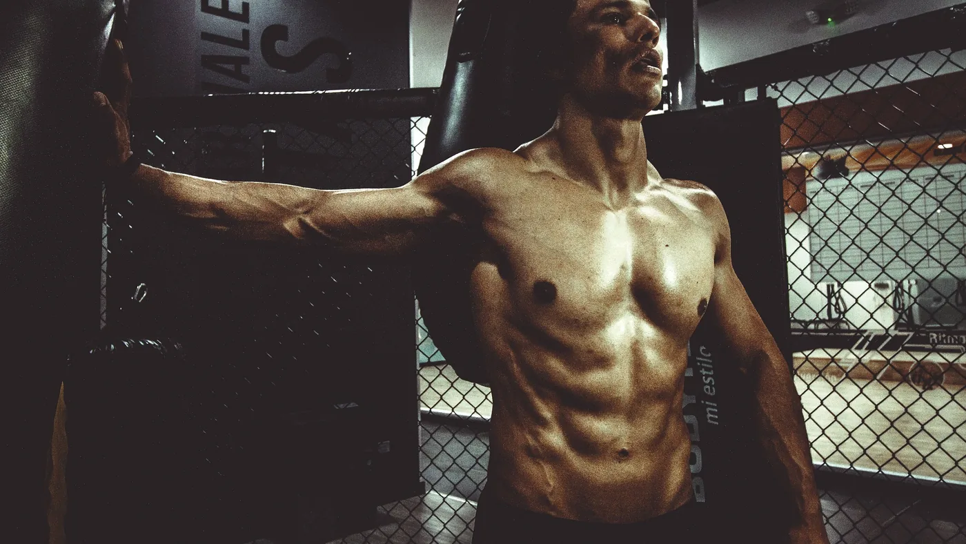 Shirtless man inside an MMA cage leans on the fence in a dim gym, with a focused, intense expression.