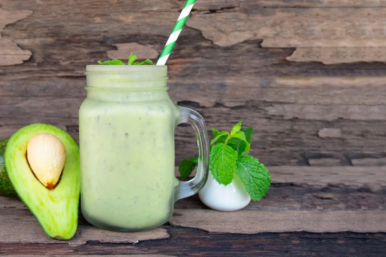 Avocado smoothie in a mason jar with striped straw, beside a halved avocado and mint on rustic wood.