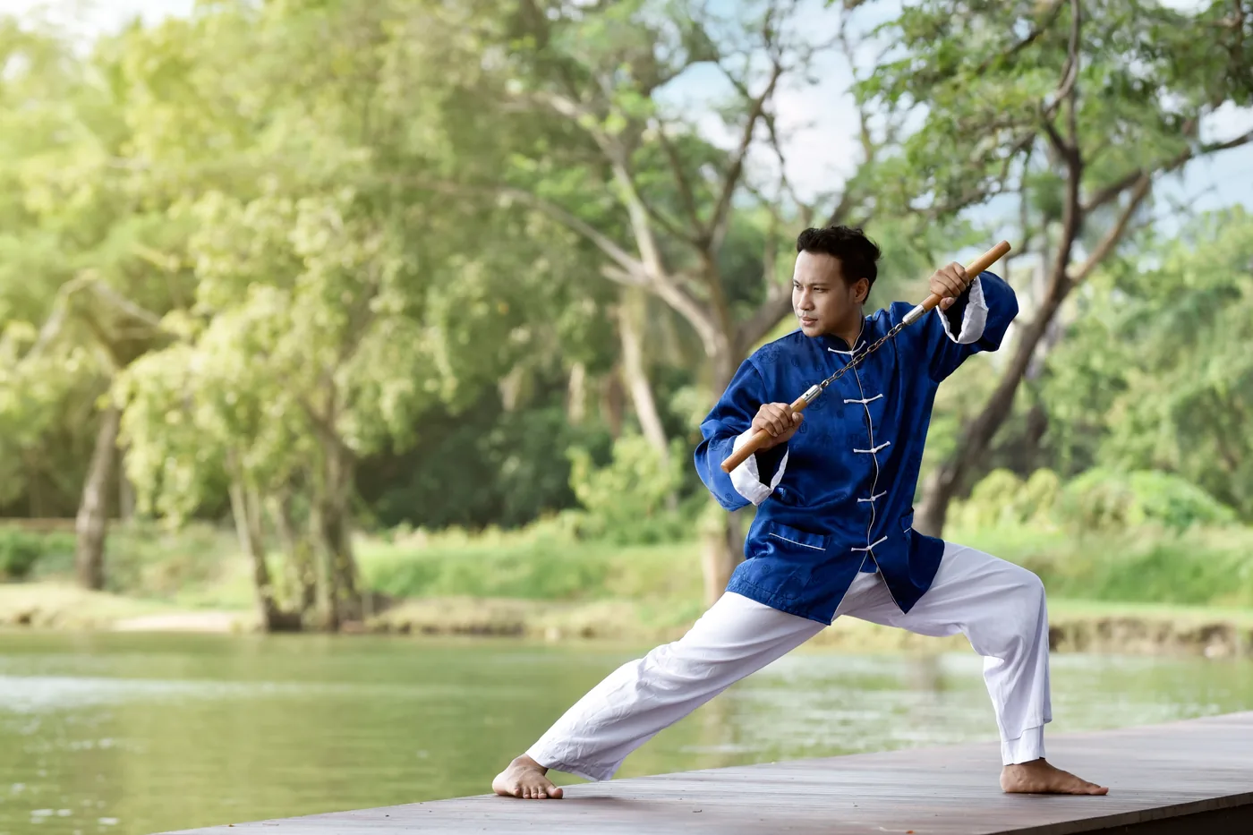 Man in blue martial arts uniform practices nunchucks on a wooden dock by a calm lake with green trees.