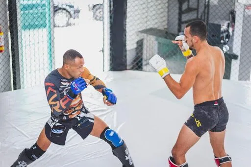 Two MMA fighters spar on a white mat in a gym, one shirtless and one in a black patterned rash guard, both in guard stance.
