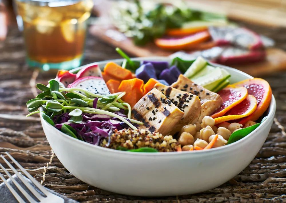 Colorful grain bowl with grilled tofu, chickpeas, sprouts, and fruit on a rustic table, with a drink in the background.