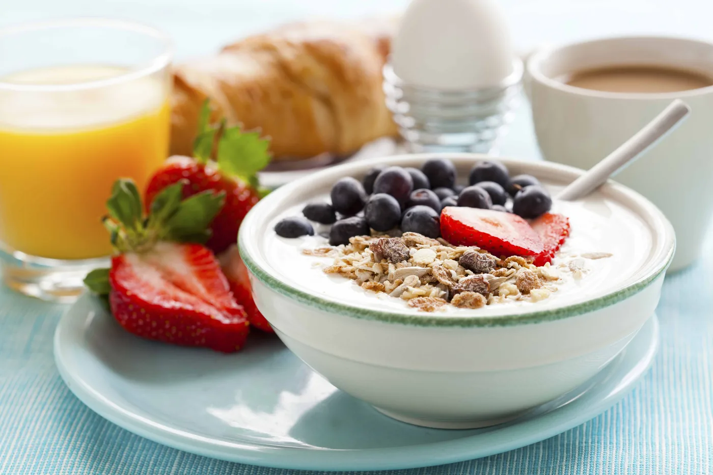 Bowl of yogurt topped with granola, blueberries and strawberries, with orange juice, coffee and croissant on a bright table.