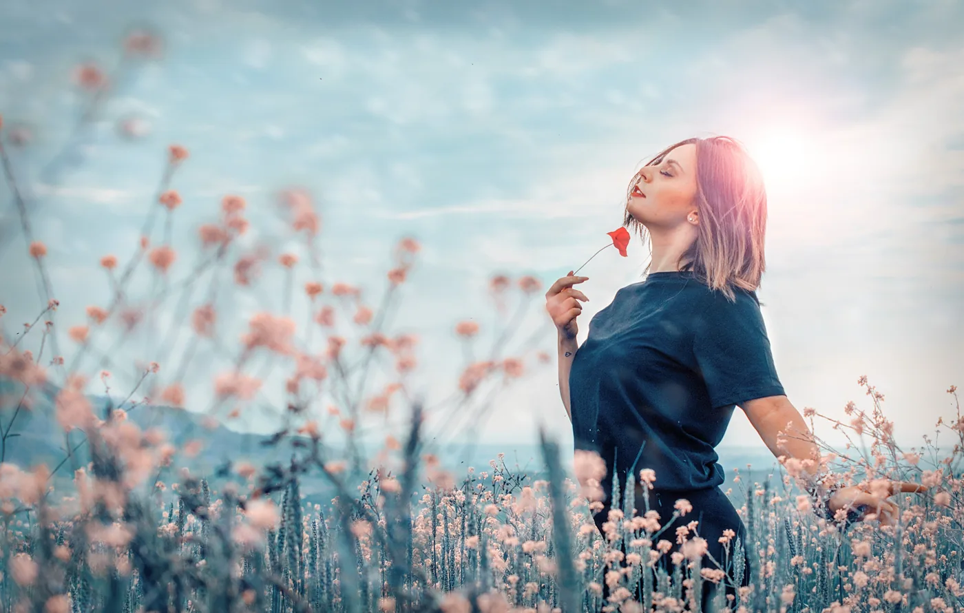 Woman in a pink wildflower field holds a red flower under a bright blue sky, bathed in dreamy sunlight.