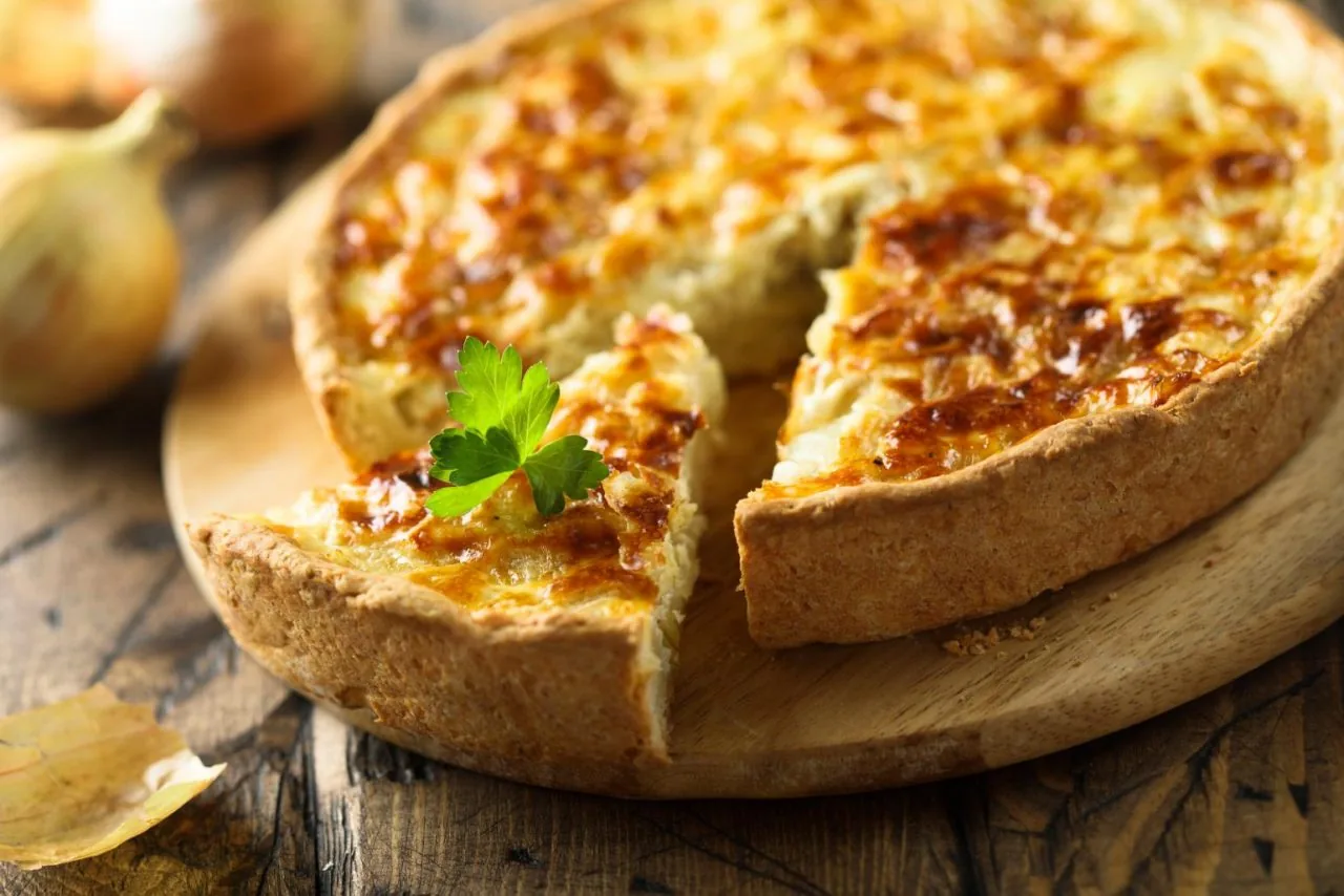 Golden quiche on a wooden board with a slice cut out, topped with parsley, on a rustic table.