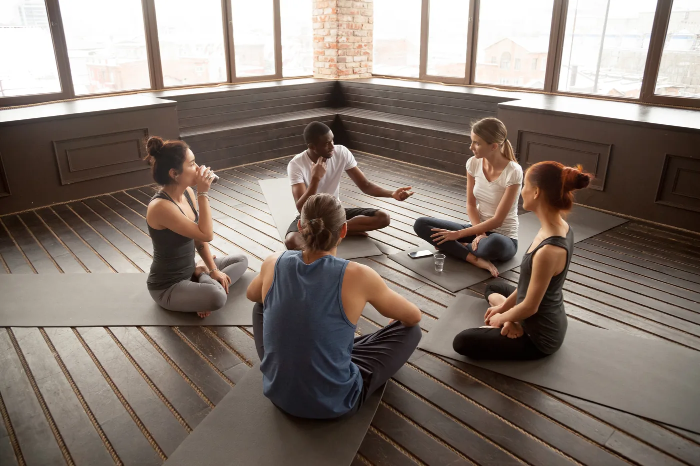Five people sit cross-legged on yoga mats in a bright studio, chatting after class near large windows and a brick pillar.