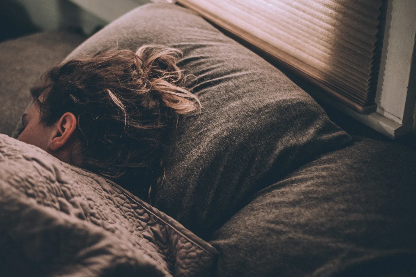 Sleeping person with tousled hair on a gray couch beside window blinds, bathed in warm sunlight.