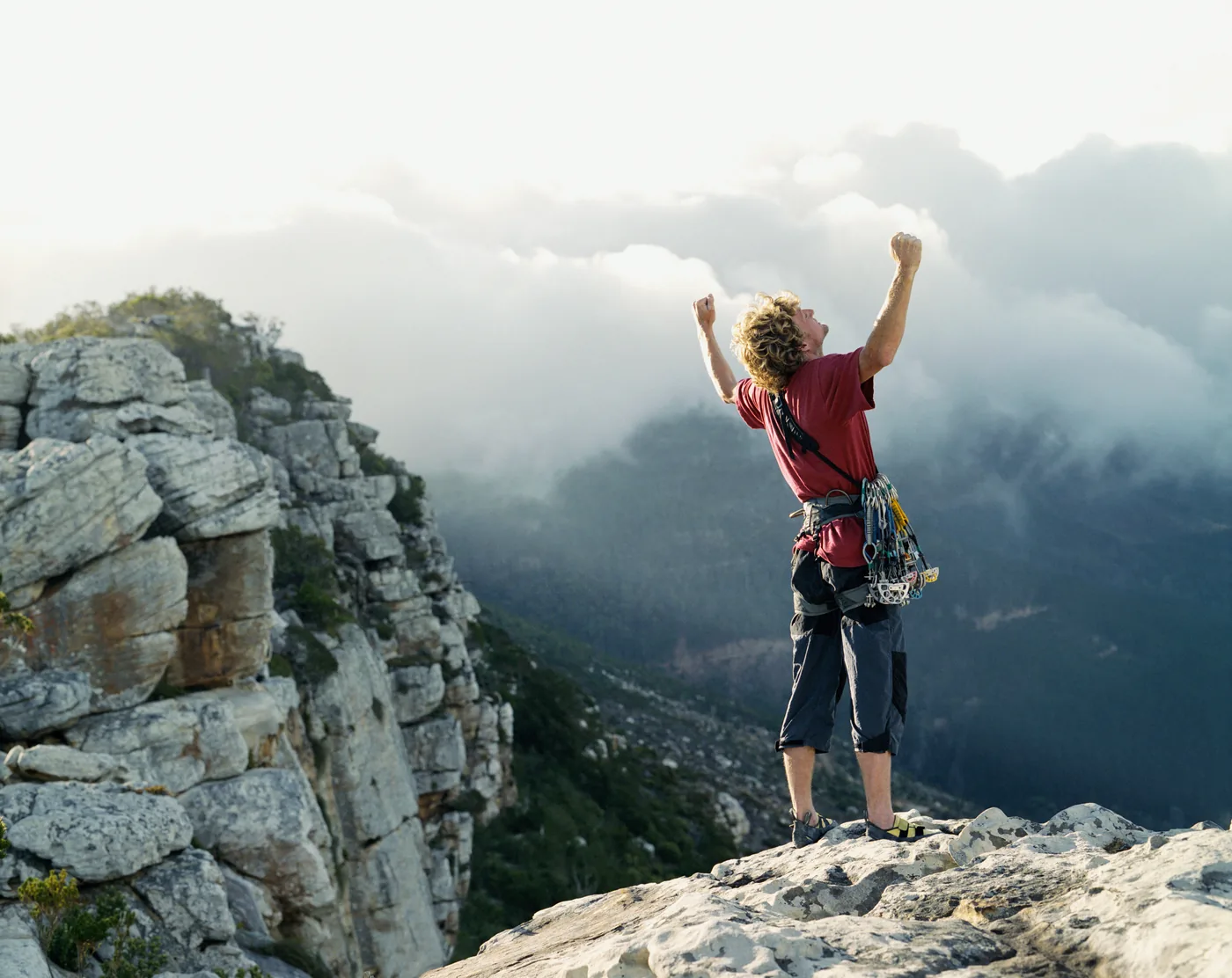 Climber in red shirt stands on rocky mountain ledge, arms raised in triumph, with misty peaks and clouds behind him.