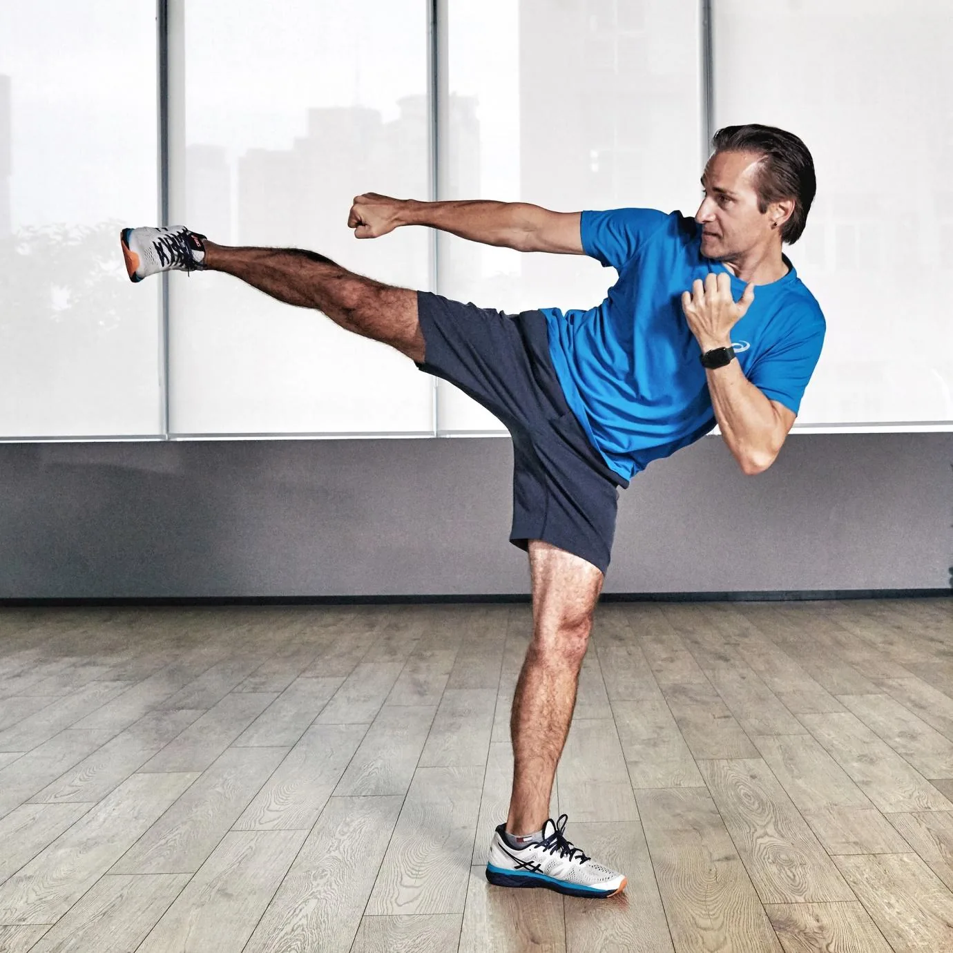 Man in blue shirt and black shorts throws a high side kick in a bright studio with wood floor and large windows.