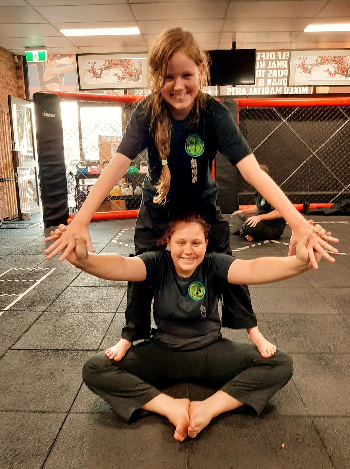 Two smiling girls in black martial arts uniforms pose playfully in a gym with red cage walls and mirrored wall text.