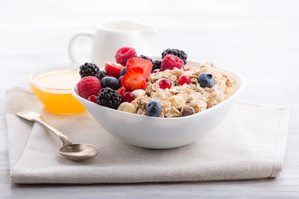 Bowl of oatmeal topped with berries on a napkin, with honey, spoon, and white mug in a bright breakfast setting