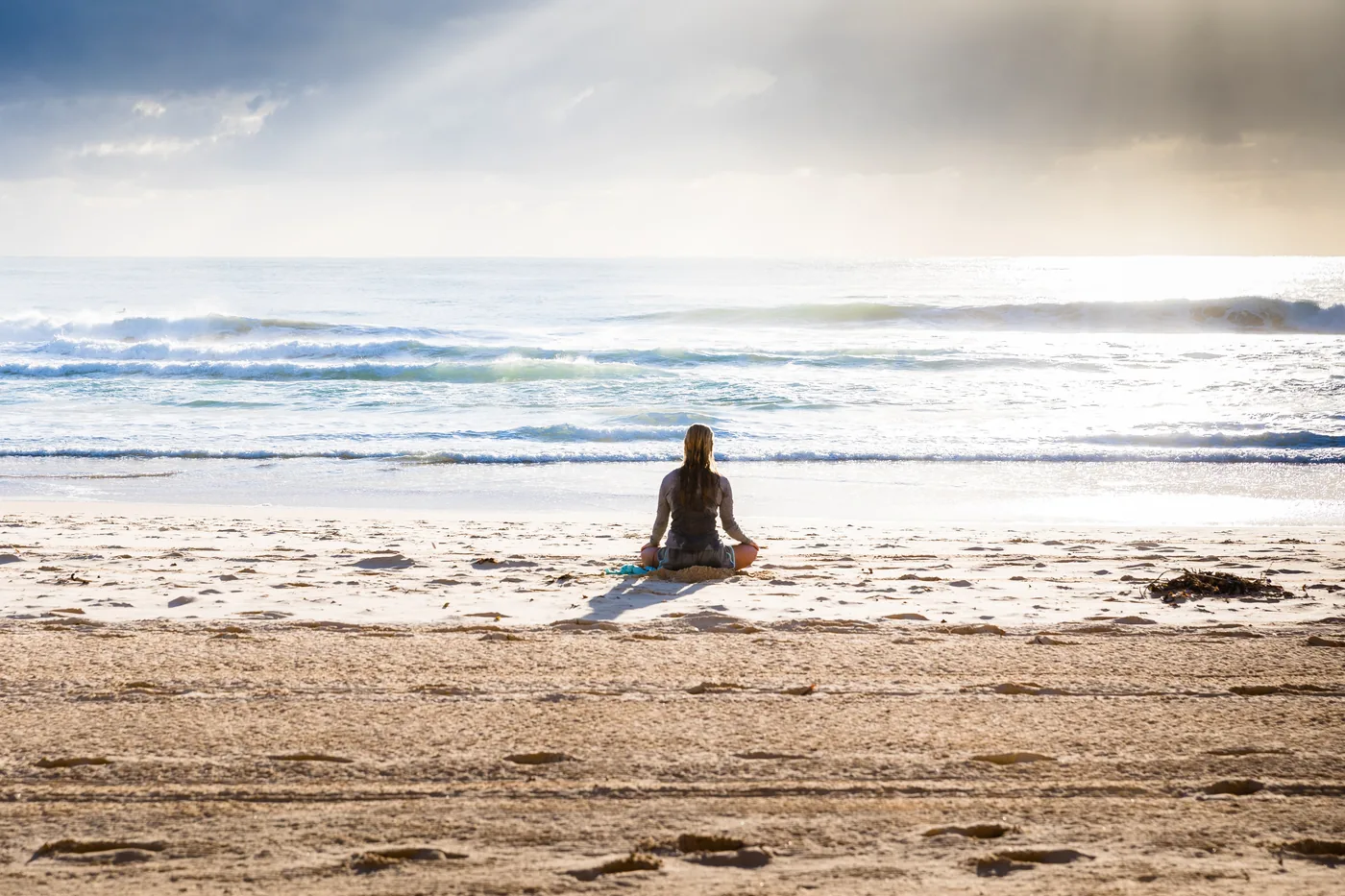 Woman meditating cross-legged on a sunny beach, facing calm ocean waves under a bright cloudy sky.