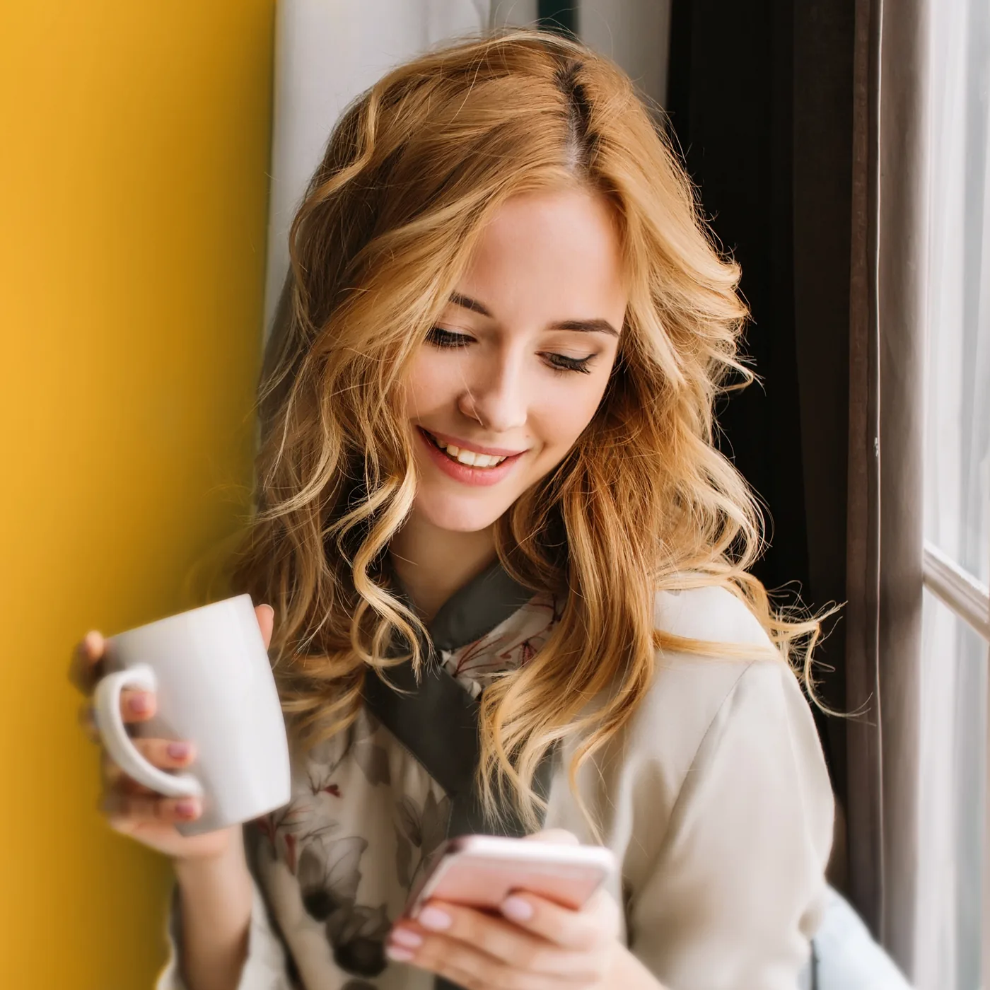Smiling blonde woman by a window holding a white mug and looking at her phone, in a warm, cozy indoor scene