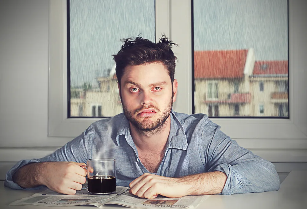 Tired man sits at a table holding coffee over a newspaper, looking at camera in front of a rainy window and blurred buildings.