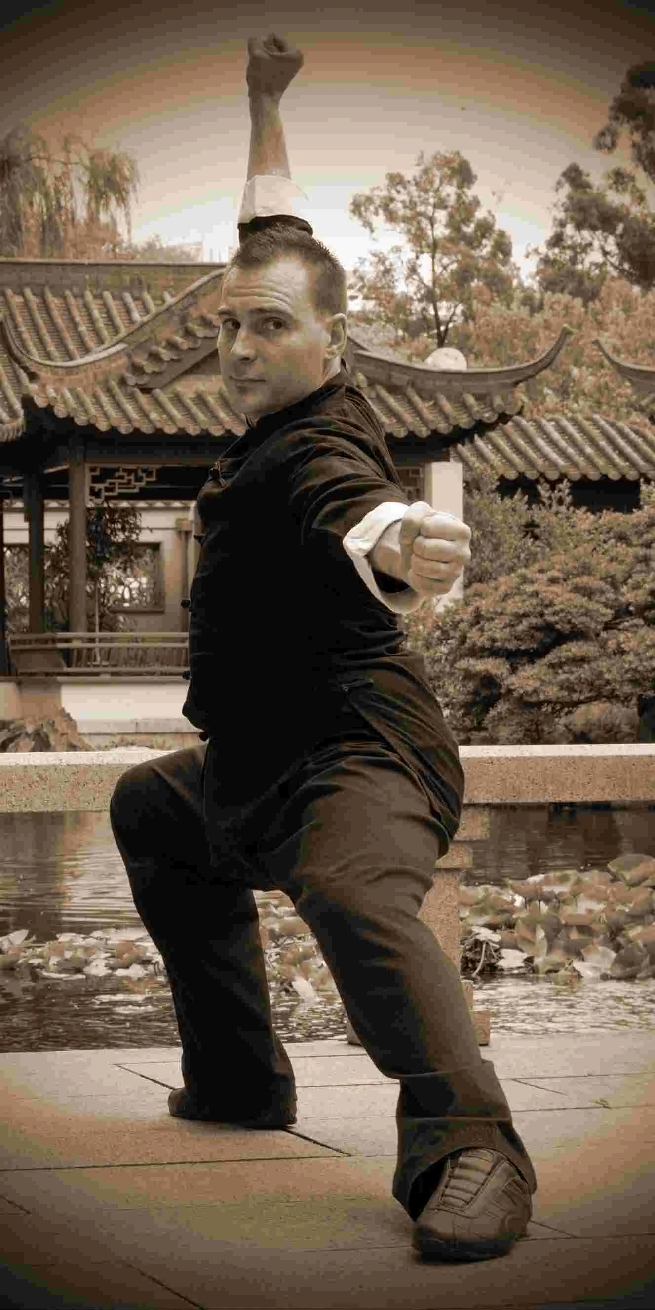 Man in black martial arts outfit strikes a kung fu pose in a traditional garden pavilion, with a focused expression.