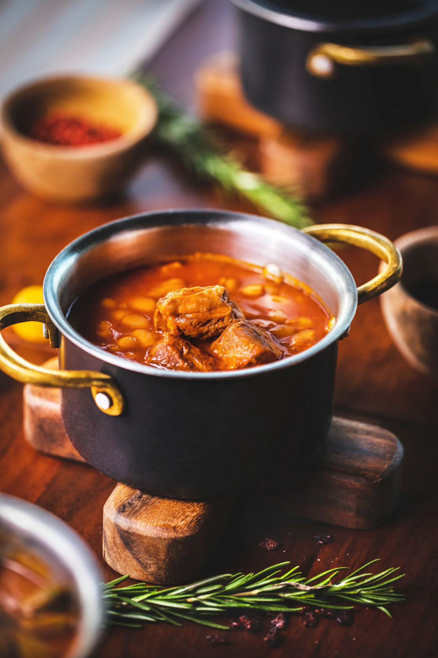 Black pot of beef and bean stew on a wooden board with rosemary sprigs, in a warm rustic food styling scene.