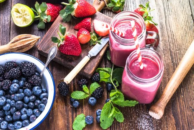 Two berry smoothies in mason jars on a rustic table with strawberries, blueberries, blackberries, mint, and a bowl of berries