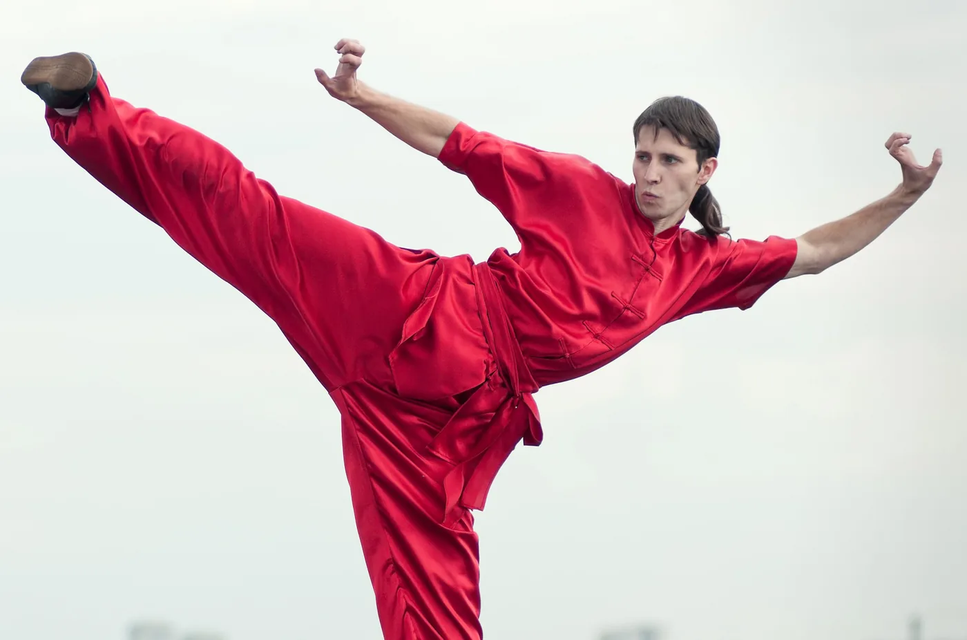 Martial artist in a red satin uniform performs a high kick pose against a bright, empty sky, looking focused.
