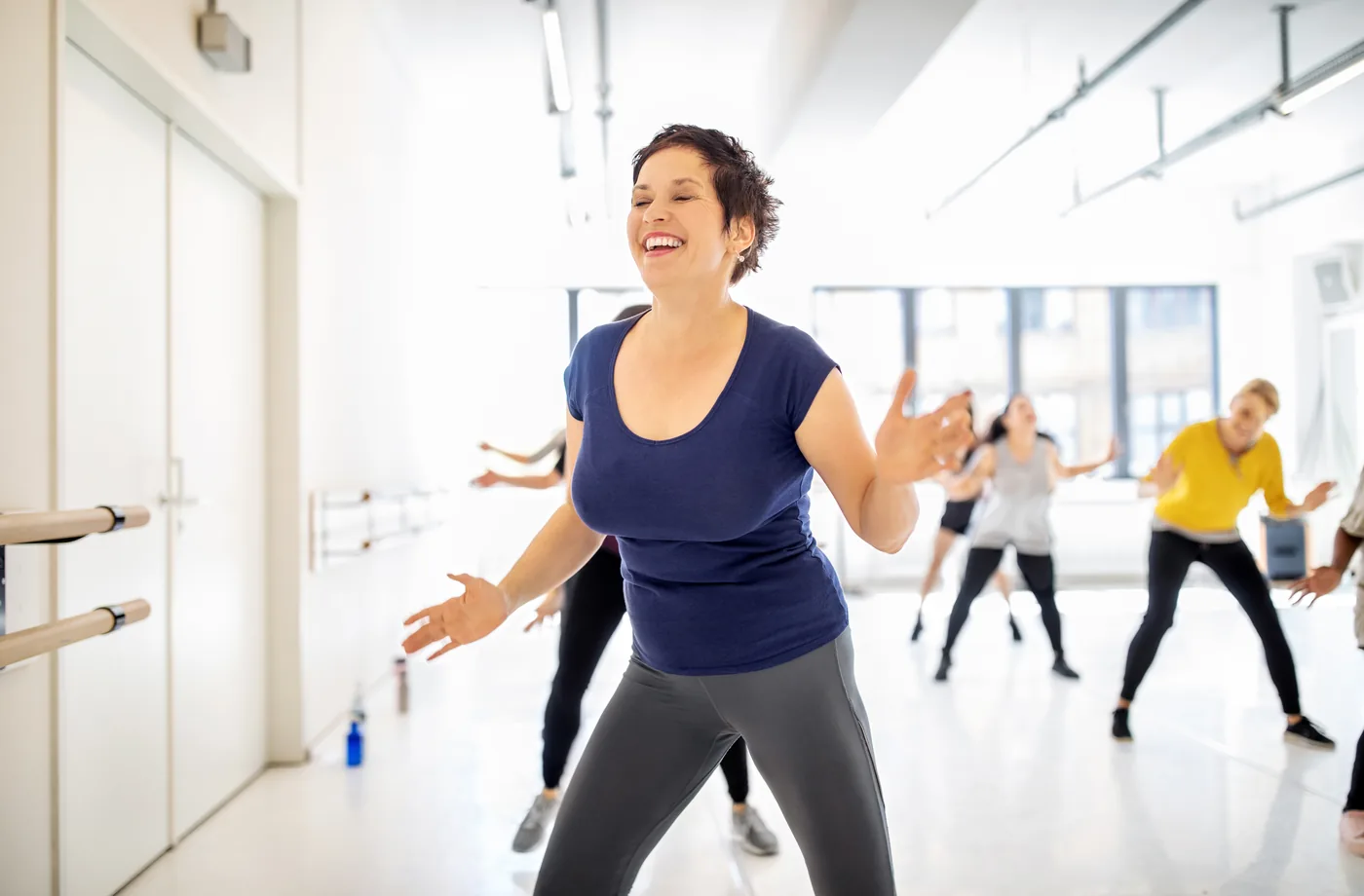 Smiling woman leads a dance workout class in a bright studio, with other participants moving behind her.