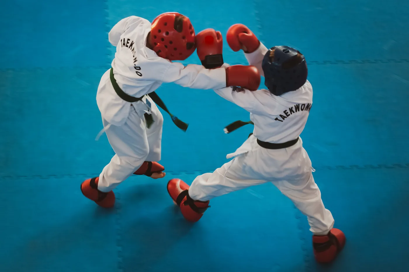 Two taekwondo kids spar on a blue mat, wearing white uniforms, red gloves and helmets; TAEKWONDO text on backs.