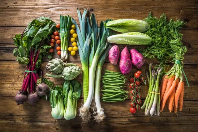 Assorted fresh vegetables—beets, leeks, lettuce, carrots, green beans, artichokes and herbs—arranged on a rustic wooden table.