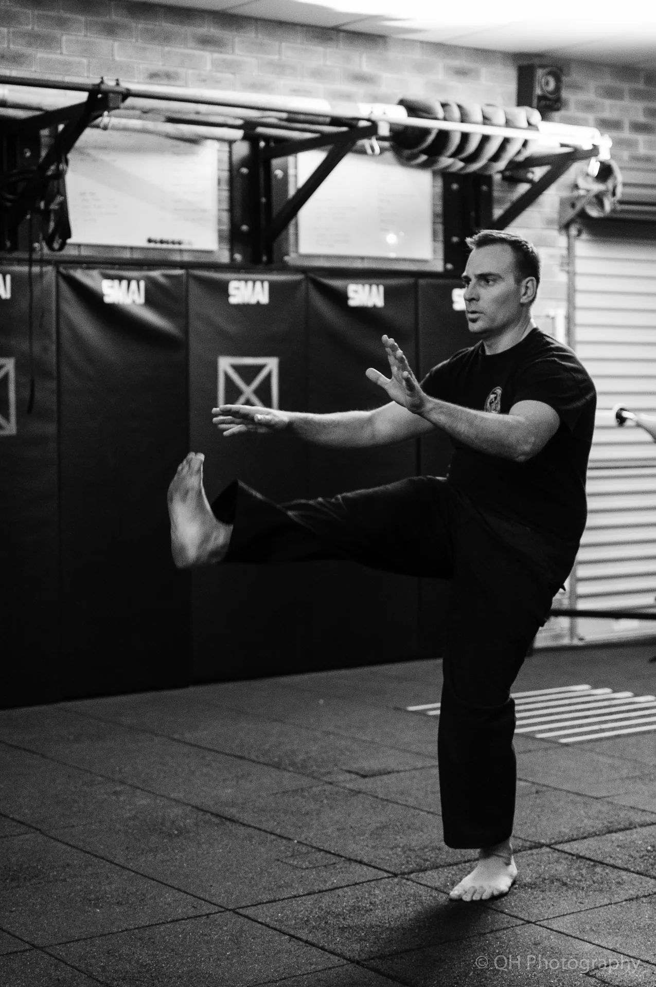 Barefoot man practices a high leg swing for dynamic flexibility in a martial arts gym with padded walls labeled SMAI, black-and-white, focused mood
