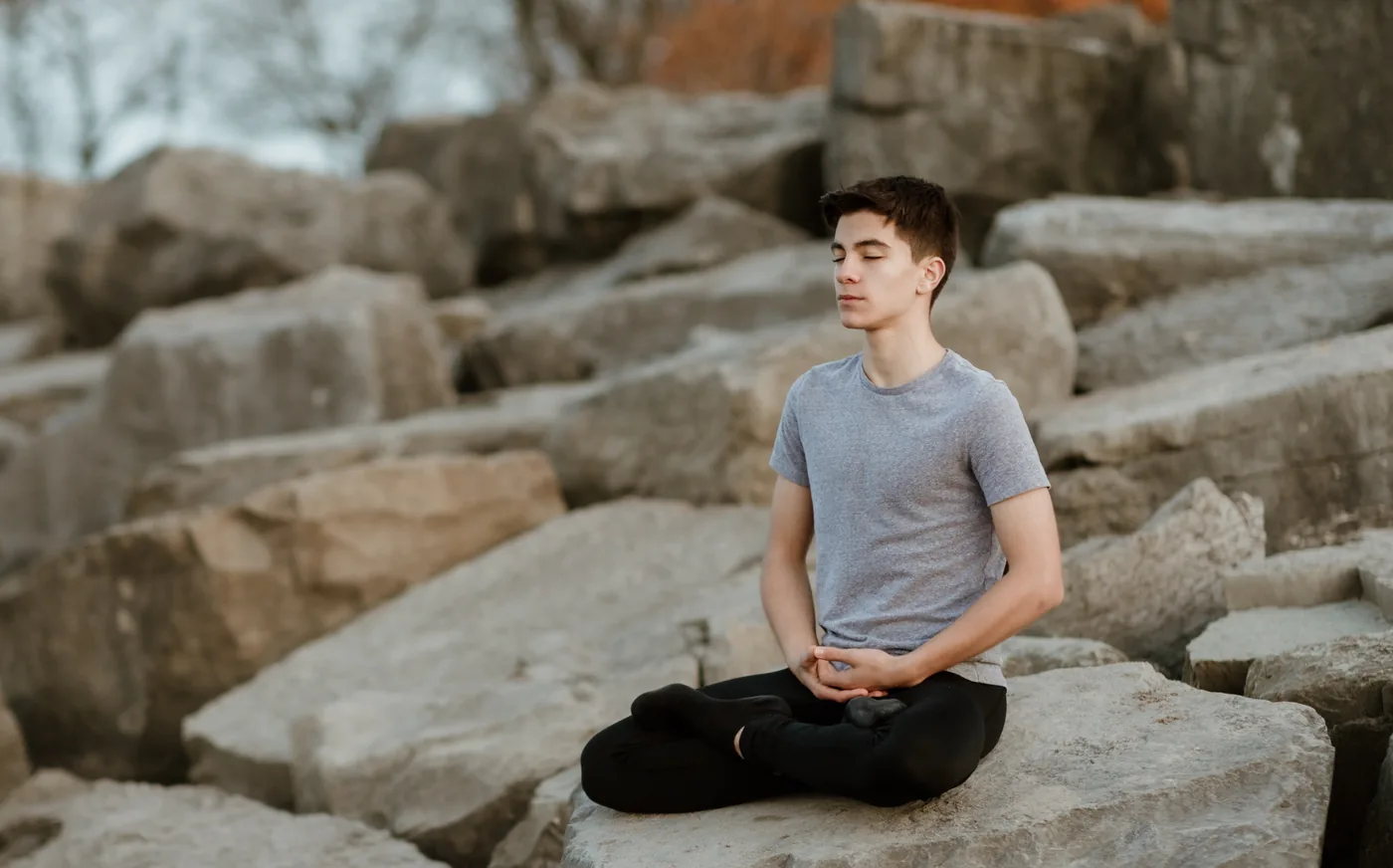 Young man meditates cross-legged on large rocks outdoors, eyes closed, calm and peaceful