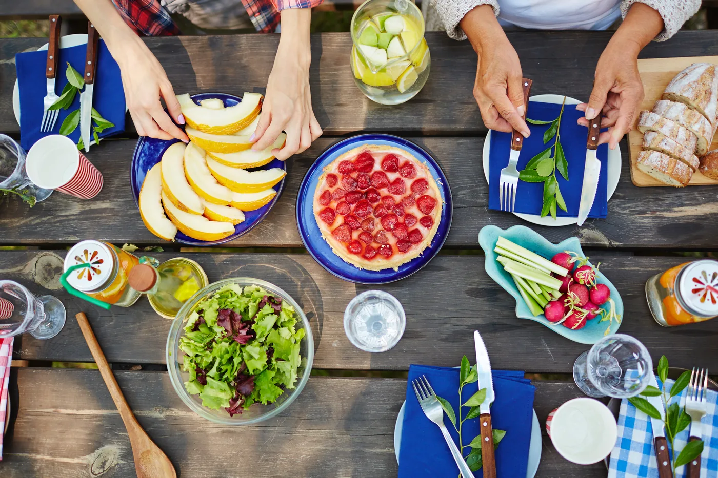 Overhead view of a shared outdoor meal with hands serving melon, fruit tart, salad, bread, radishes, and blue napkins on a rustic table