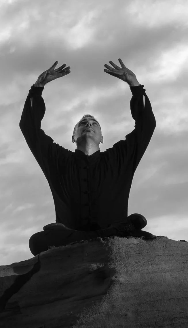 Black-and-white photo of a man sitting cross-legged on a rock, arms raised toward a cloudy sky in a calm, meditative pose.