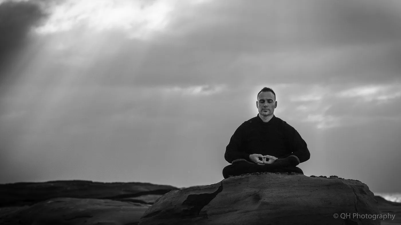 Black-and-white photo of a man meditating cross-legged on a rock at a cloudy beach, with QH Photography watermark.