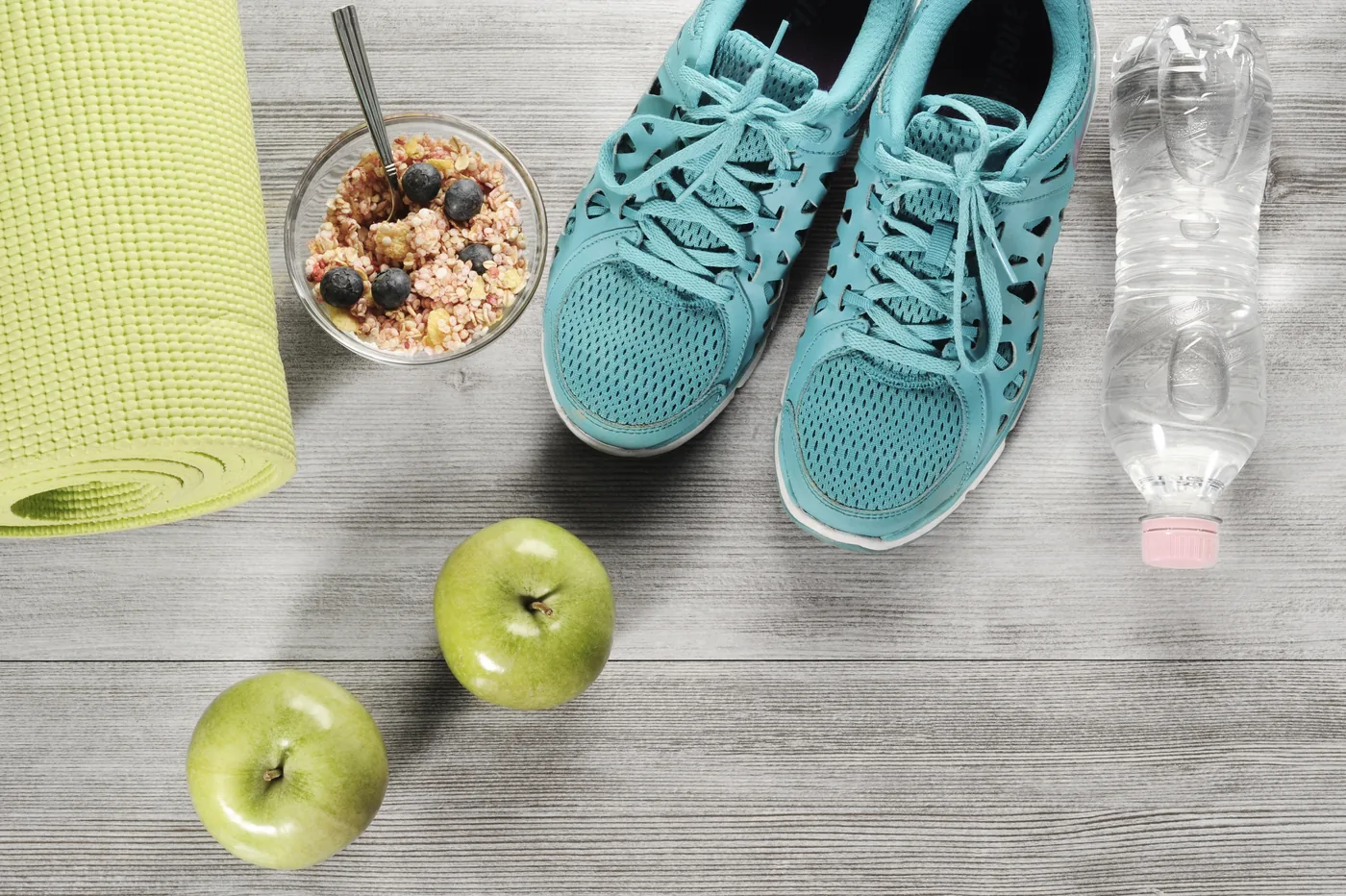 Blue running shoes, yoga mat, granola with blueberries, water bottle, and green apples on a gray wood floor.