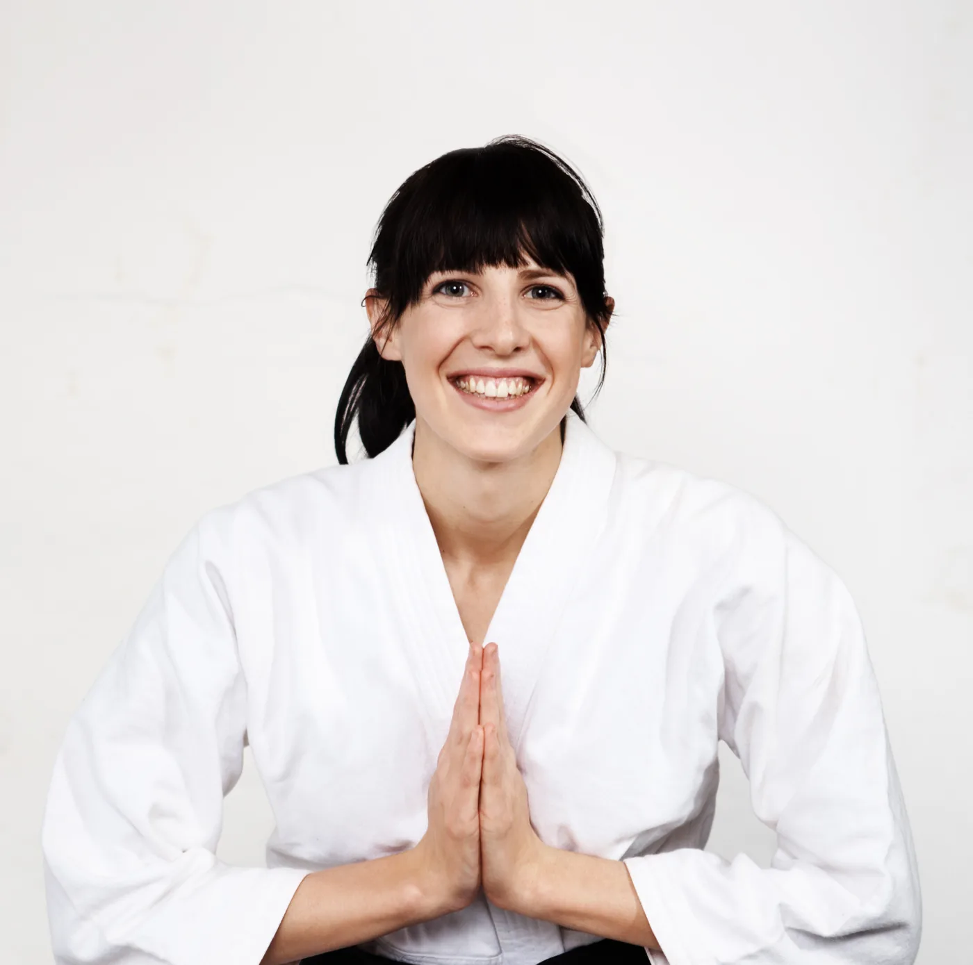 Smiling woman in a white martial arts gi holds palms together in greeting against a plain white background.