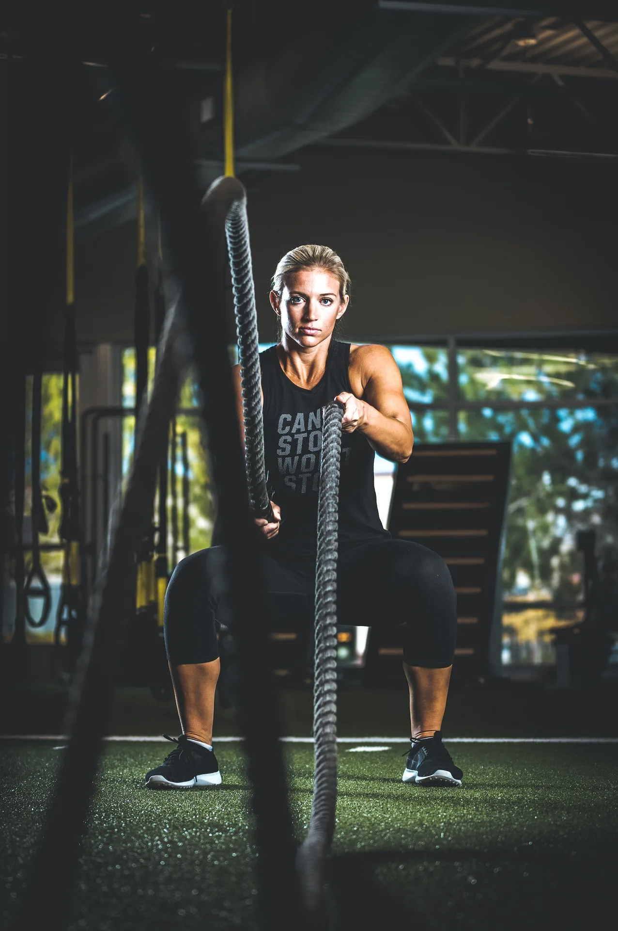 Woman in a gym does battle rope exercises, wearing black workout clothes and a focused, determined expression; shirt reads CAN'T STOP WON'T STOP
