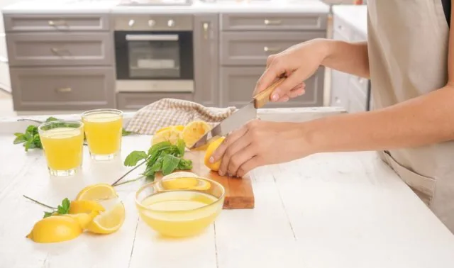 Person slices a lemon on a cutting board in a bright kitchen, surrounded by lemon drinks, mint, and lemon wedges.