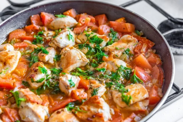 Skillet of chicken and tomatoes simmering on a stovetop, topped with fresh herbs and orange-red sauce.