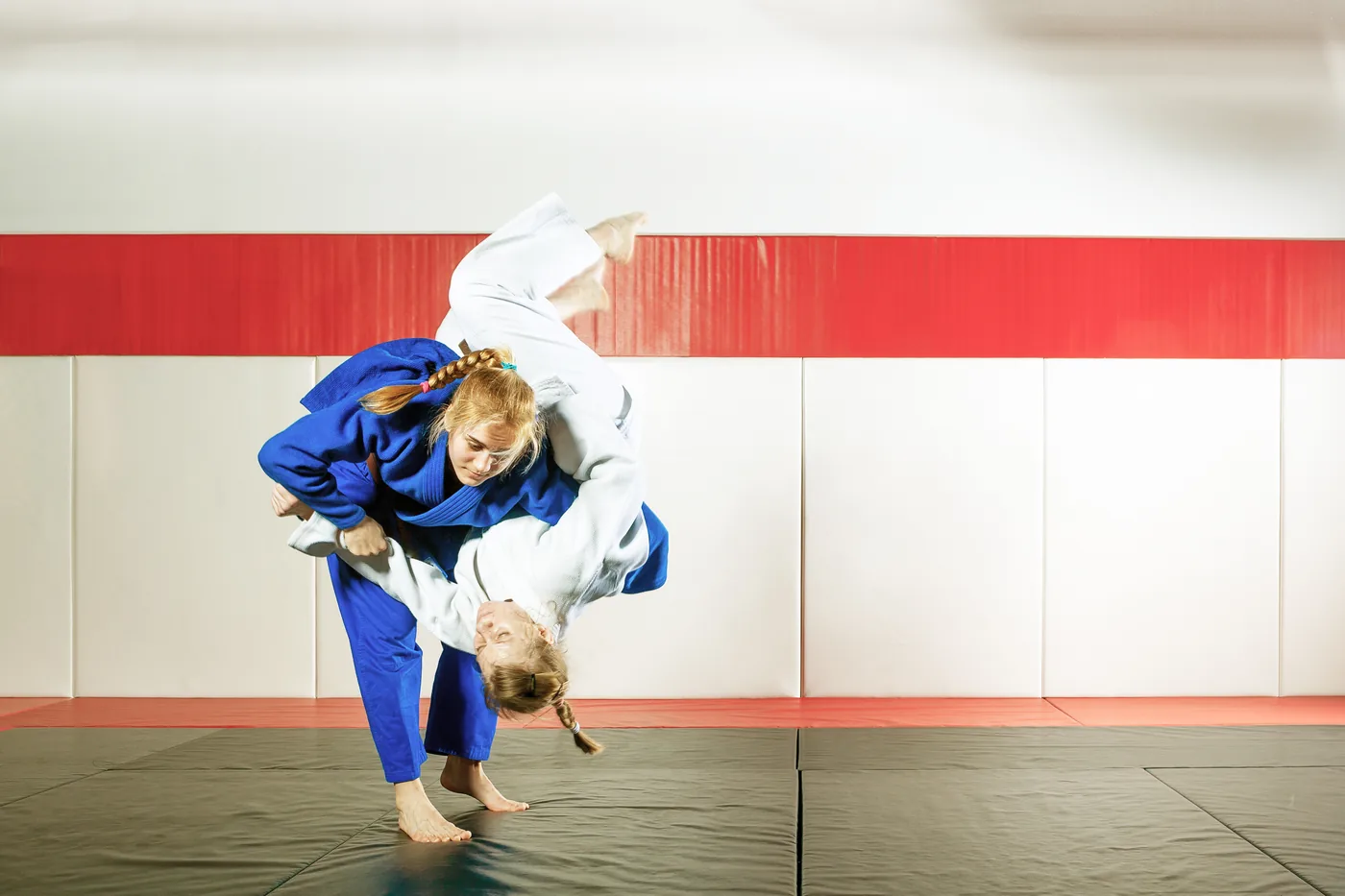 Two girls in blue and white judo gis practice a throw on tatami mats in a bright dojo with red and white wall panels.