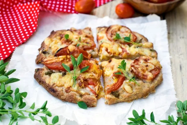 Four slices of rustic pizza with tomatoes and herbs on parchment, on a wooden table beside a red polka-dot cloth.