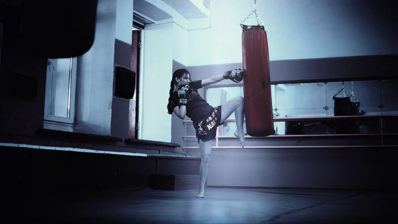 Woman in boxing gloves kicks a red heavy bag in a dim martial arts gym, with mirrors and cool blue lighting.