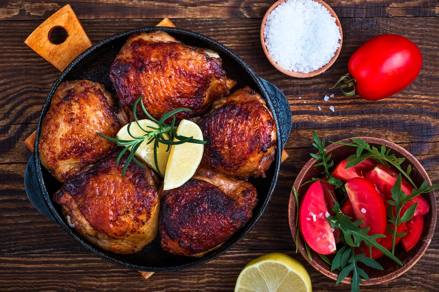 Top-down view of roasted chicken thighs in a cast-iron skillet with lemon and rosemary, beside salt and tomatoes on rustic wood.