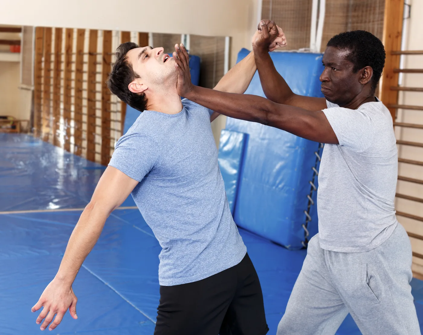 Two men practice martial arts in a gym, one using a palm strike to the others and face. They are training on blue mats, tense and focused.