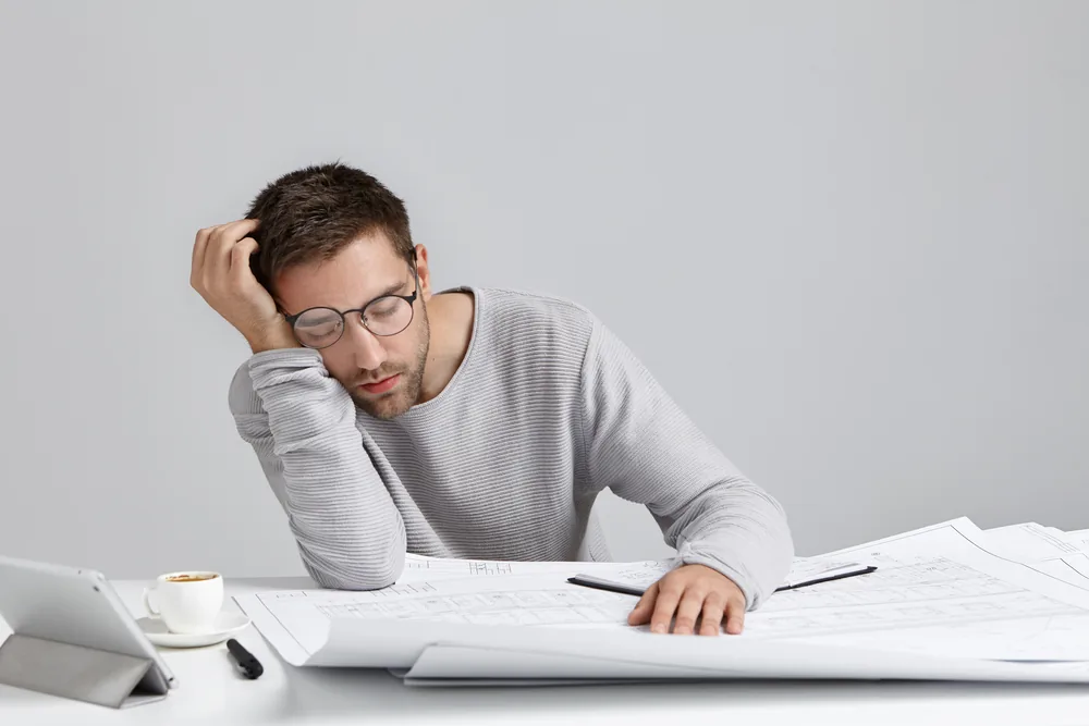 Tired man with glasses naps over architectural blueprints at a desk, with a tablet and coffee cup beside him in a gray office.