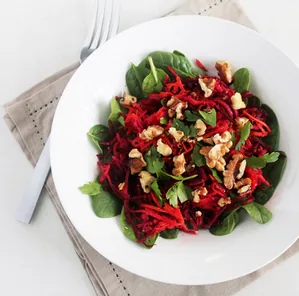 White plate of spinach salad with shredded beets, walnuts, and herbs on a napkin, with a fork on a white table.